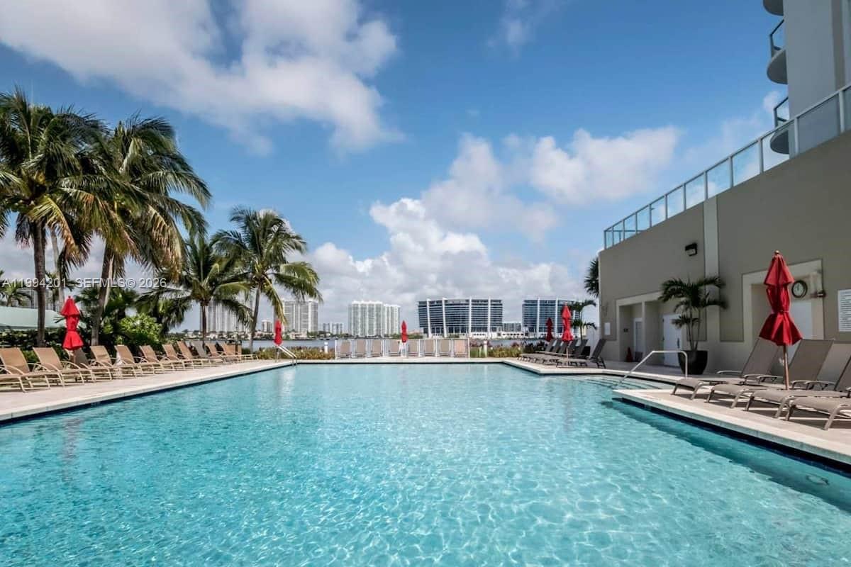 A swimming pool with lounge chairs and red umbrellas sits outdoors near a building — SUNNY ISLES BEACH, FL