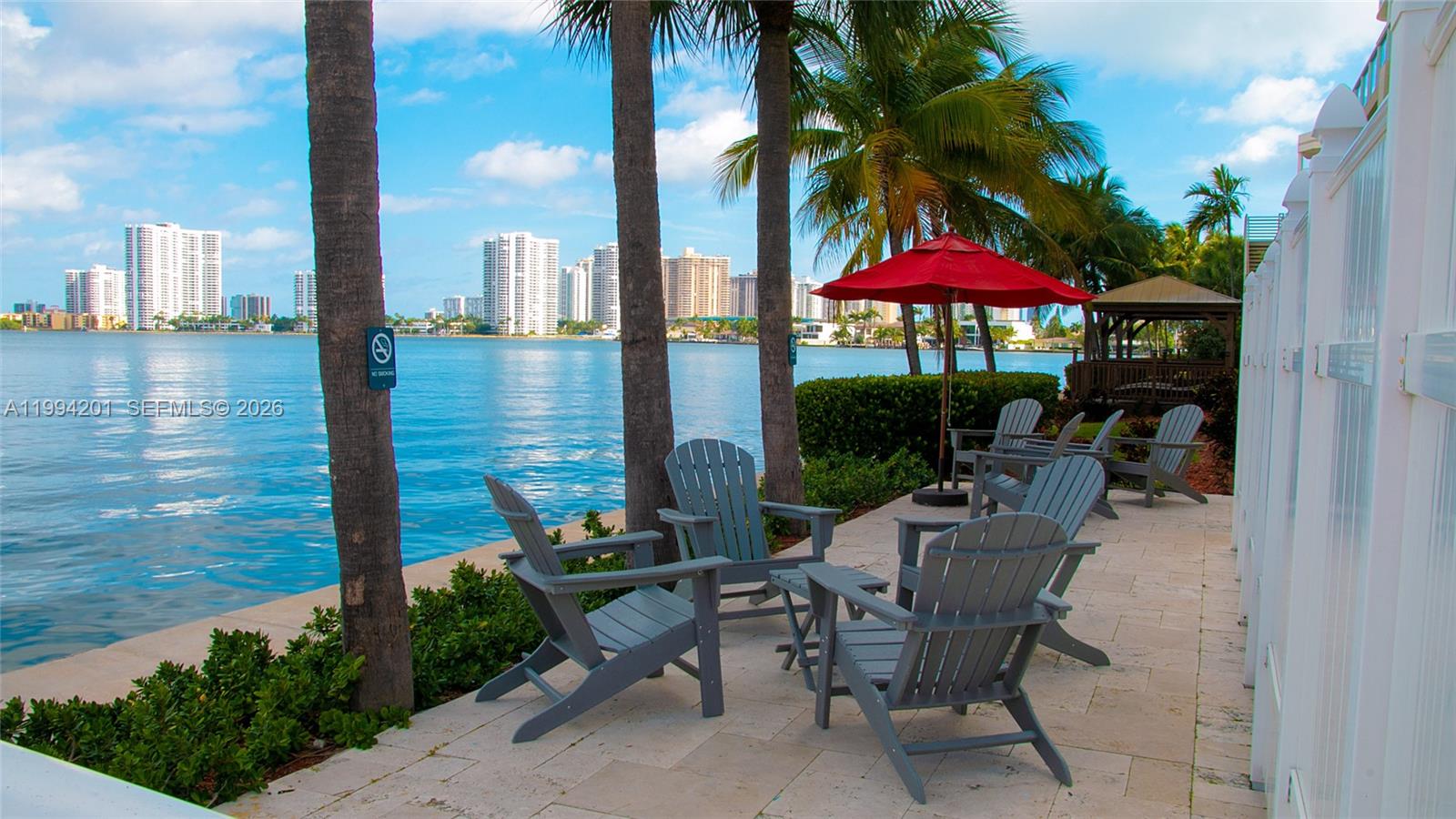 A waterfront patio features gray Adirondack chairs, a red umbrella, and palm trees — SUNNY ISLES BEACH, FL