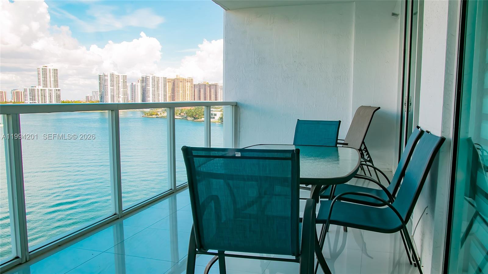 A balcony with a glass table and chairs overlooks a bay and city skyline — SUNNY ISLES BEACH, FL