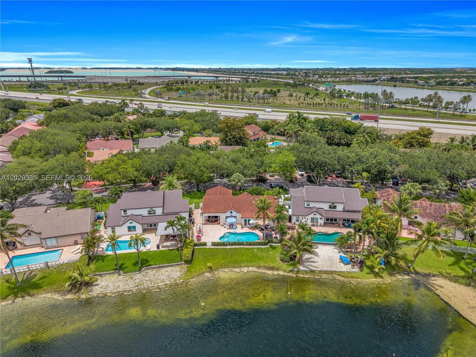 An aerial view shows three waterfront houses with swimming pools along a lake near a highway — 19641 NW 88th Ave, Hialeah FL 33018, HIALEAH, FL