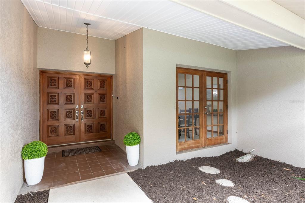 An entryway features double wooden doors, a glass-paned door, potted plants, and a tiled porch — 1056 ENISWOOD PKWY, PALM HARBOR, FL