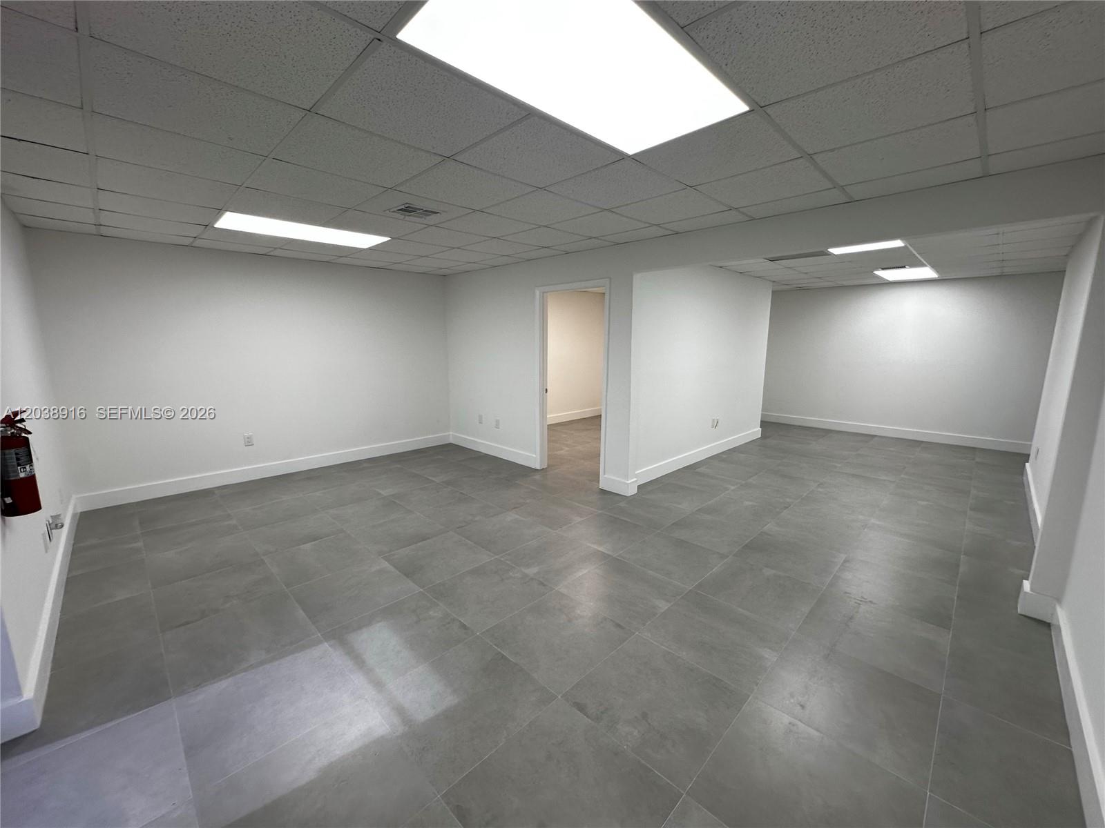 An empty room features white walls, a drop ceiling with recessed lighting, and gray tile flooring — FL