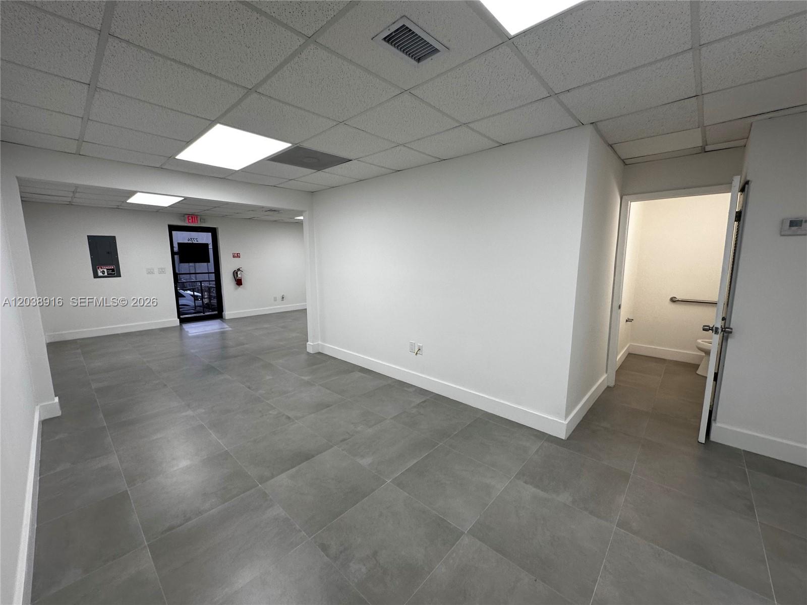 An empty office space features gray tile flooring, white walls, and a private restroom — FL