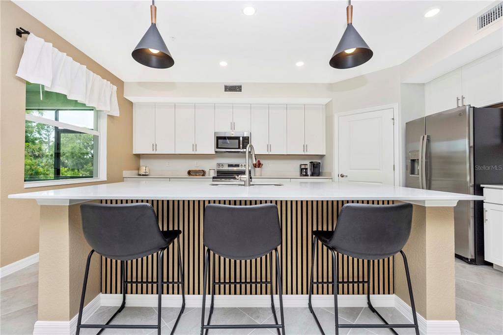 A modern kitchen features white cabinetry, a large island with bar seating, and stainless appliances — 17740 BORACAY COURT UNIT 102, VENICE, FL