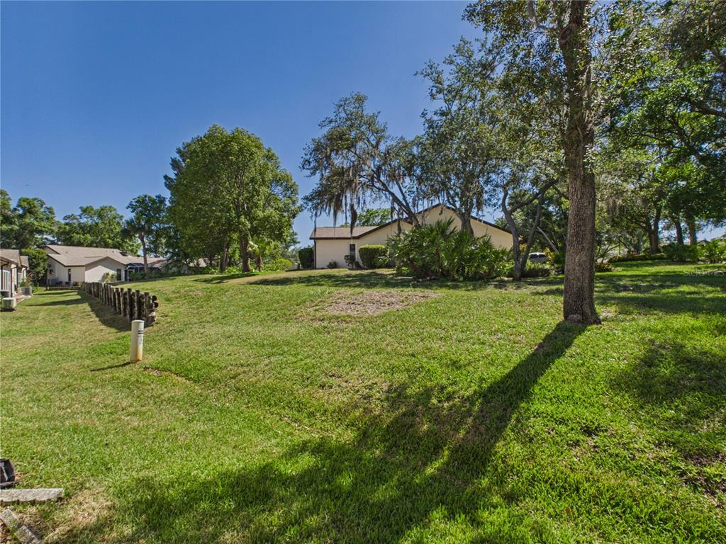 A grassy, sloped backyard features large trees and a wooden fence near a suburban house — 7511 HEATHER WALK DRIVE, WEEKI WACHEE, FL
