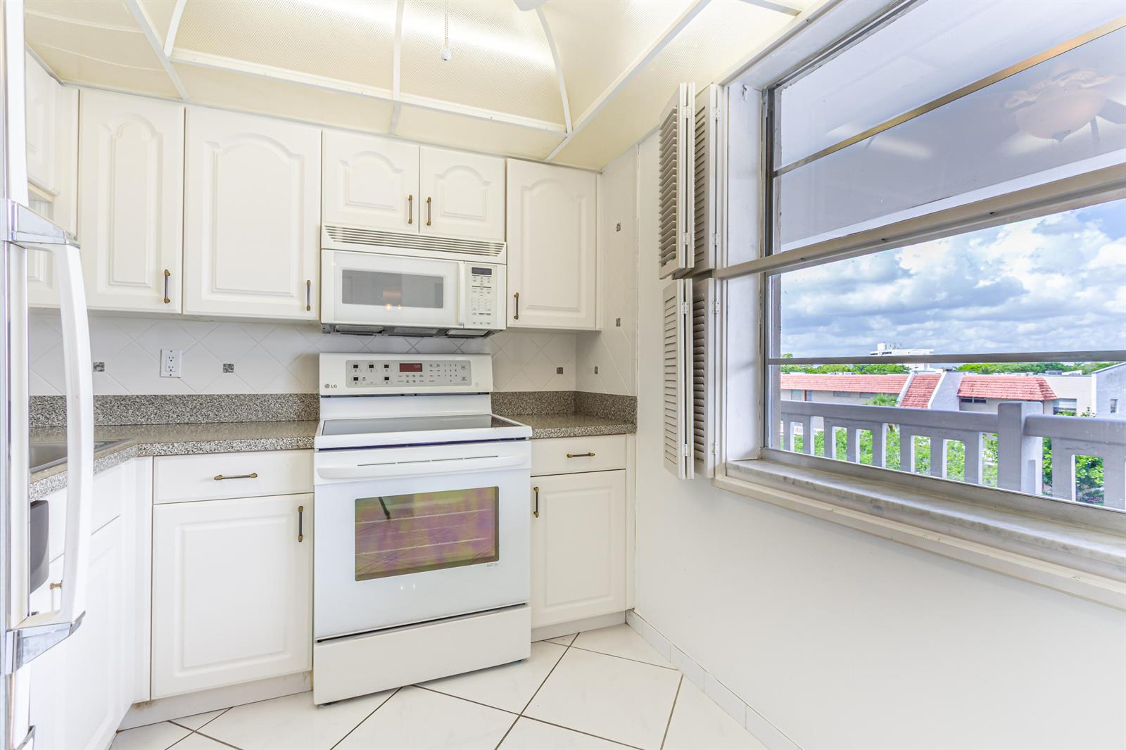 A kitchen with white cabinets, white appliances, granite countertops, and a large window overlooking buildings — 3531 Inverrary Drive # H406, Lauderhill FL 33319, LAUDERHILL, FL