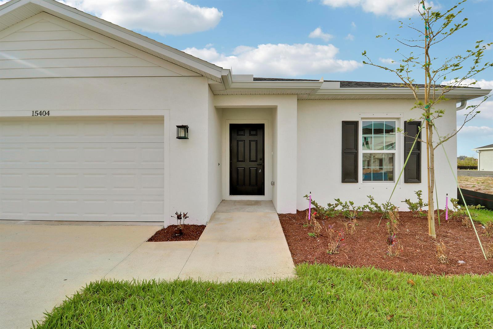 A white single-story house features a black front door, a garage, and a small garden — 15404 SW American Street, FL 34956, FL
