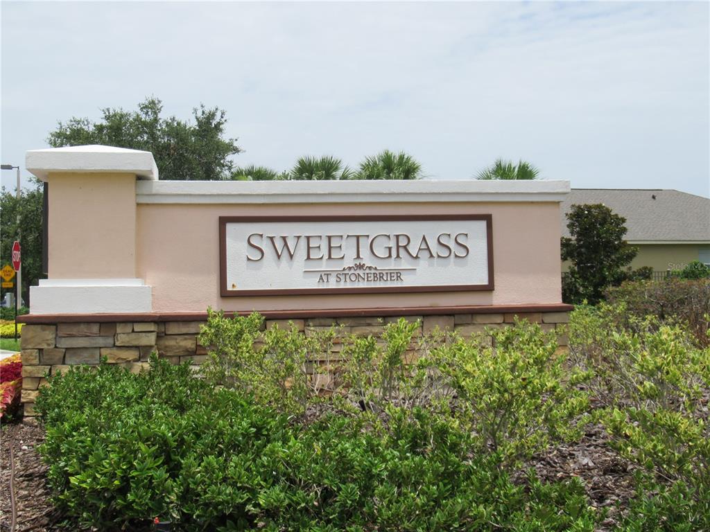 A beige stucco monument sign for Sweetgrass at Stonebrier sits above a stone base — 2822 MAPLE BROOK LOOP, LUTZ, FL