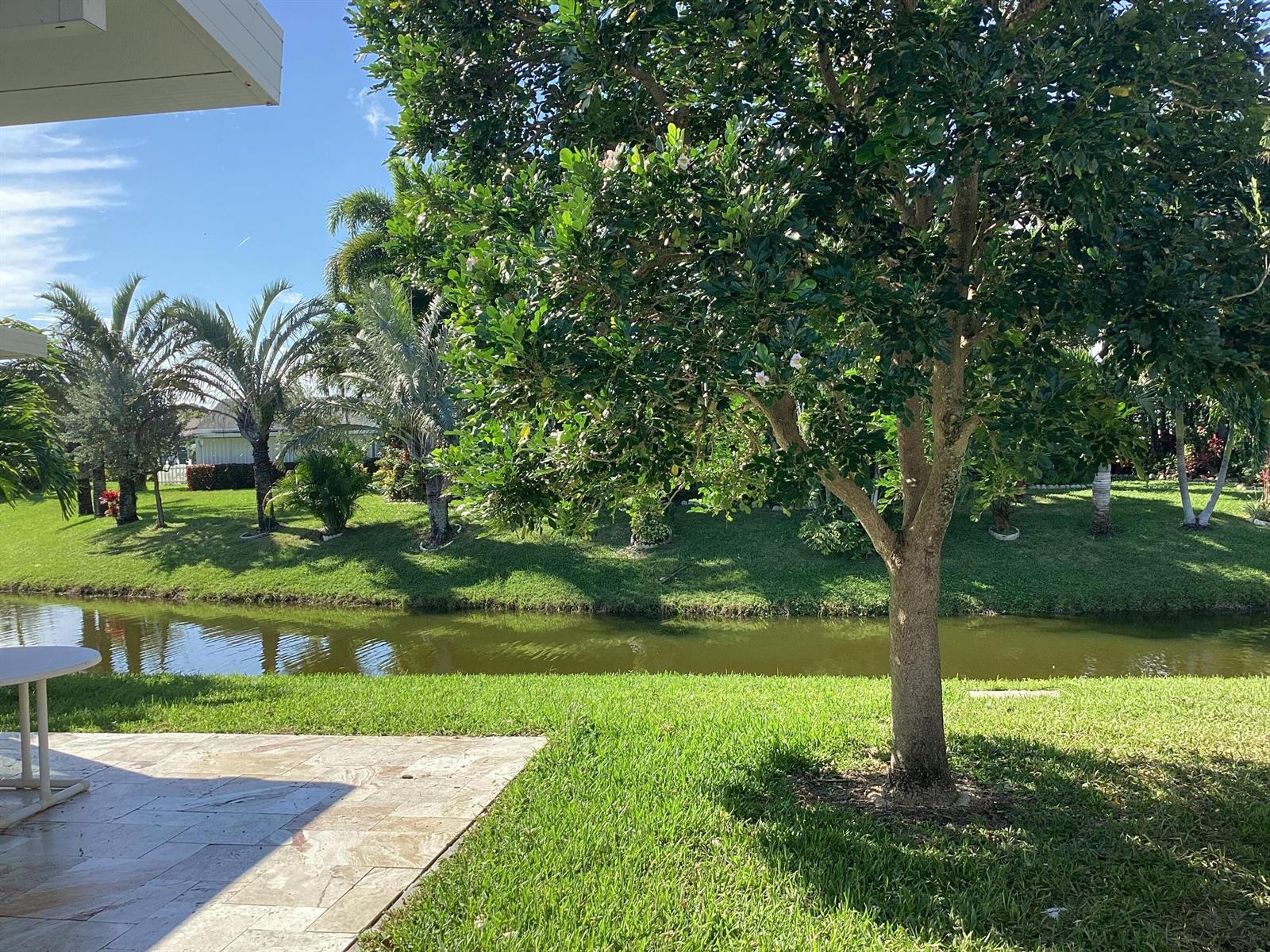 A patio overlooks a grassy lawn, a small canal, and various trees under a blue sky — 13861 Packard Terrace, Delray Beach FL 33484, DELRAY BEACH, FL