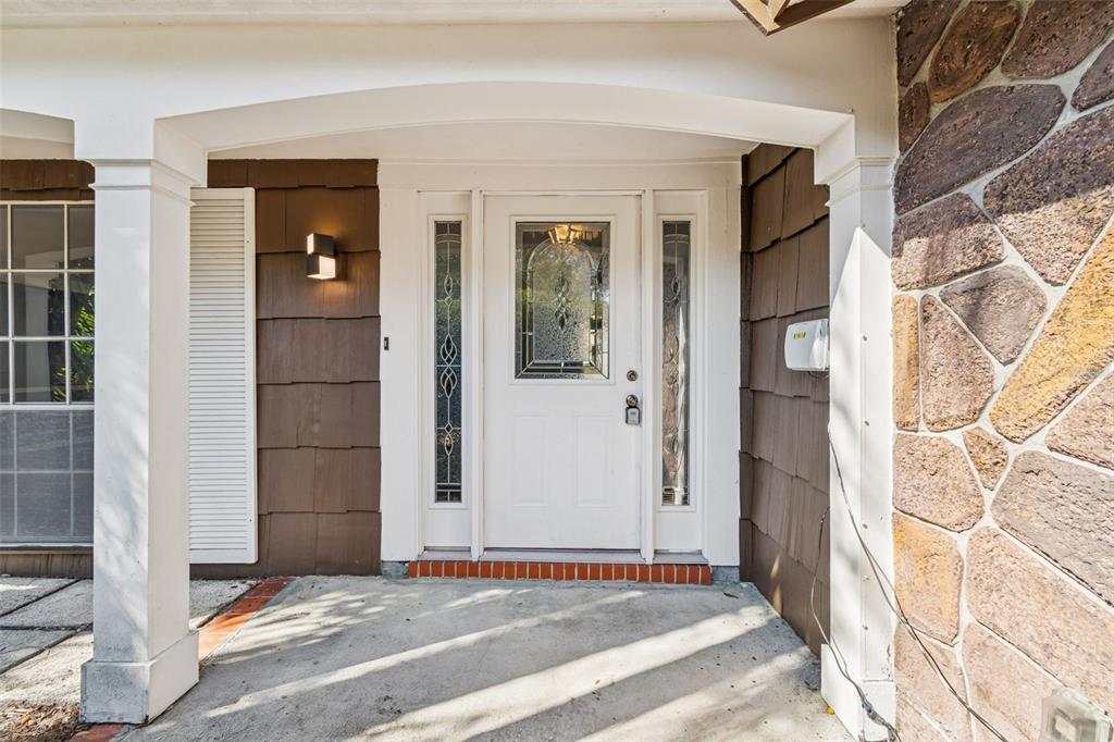 A white front door with glass inserts sits under a covered porch with stone siding — 8210 CLOVER HILL LOOP, HUDSON, FL