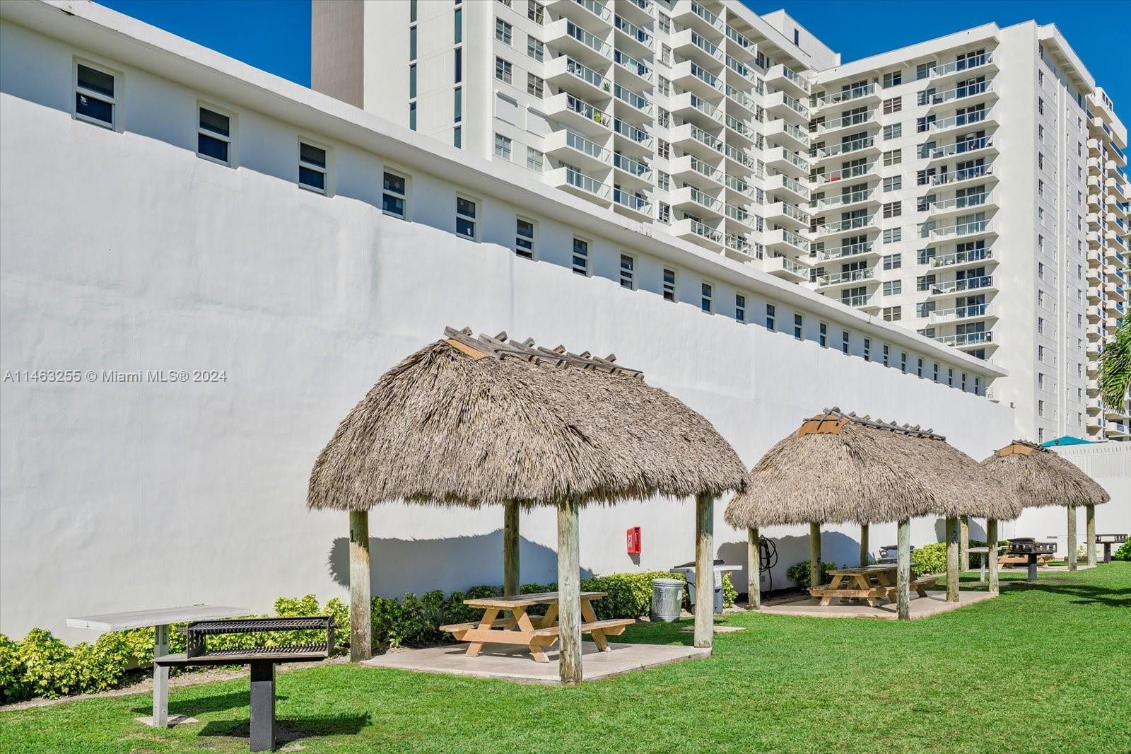 A row of thatched-roof tiki huts with picnic tables sits on a lawn beside a building — 5601 Collins Ave # 1514, Miami Beach FL 33140, MIAMI BEACH, FL