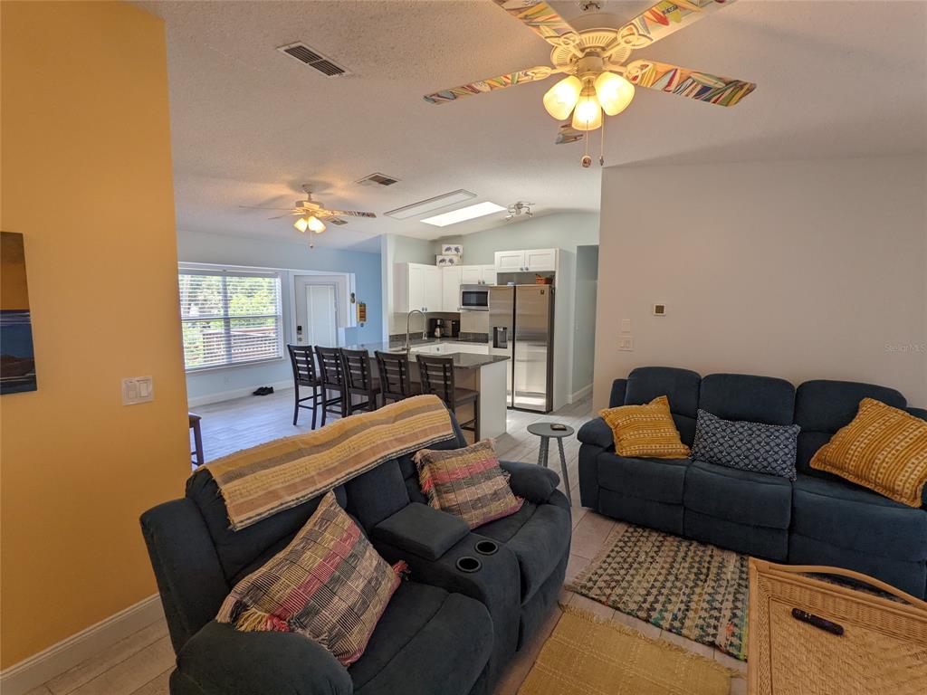 A living room with blue sofas opens into a kitchen featuring white cabinets and bar seating — 203 CANOVA DRIVE, NEW SMYRNA BEACH, FL