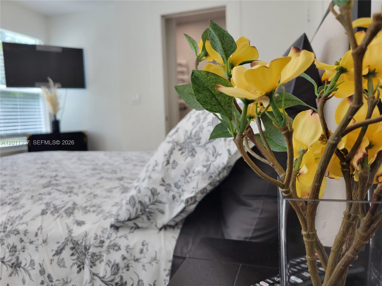 A bedroom features a bed with floral bedding, a television, and yellow artificial flowers — 735 Tradewind Dr, North Palm Beach FL 33408, NORTH PALM BEACH, FL