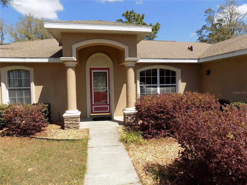 The exterior of a house features a tan facade, a covered entryway, and red-framed door — 2047 W PARAGON LANE, CITRUS SPRINGS, FL