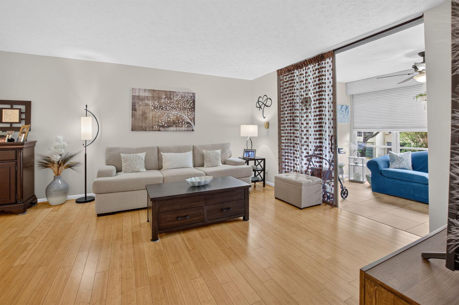 A living room with light wood floors, a beige sofa, and a sunroom beyond curtains — 11 Willowbrook Lane # 204, Delray Beach FL 33446, DELRAY BEACH, FL