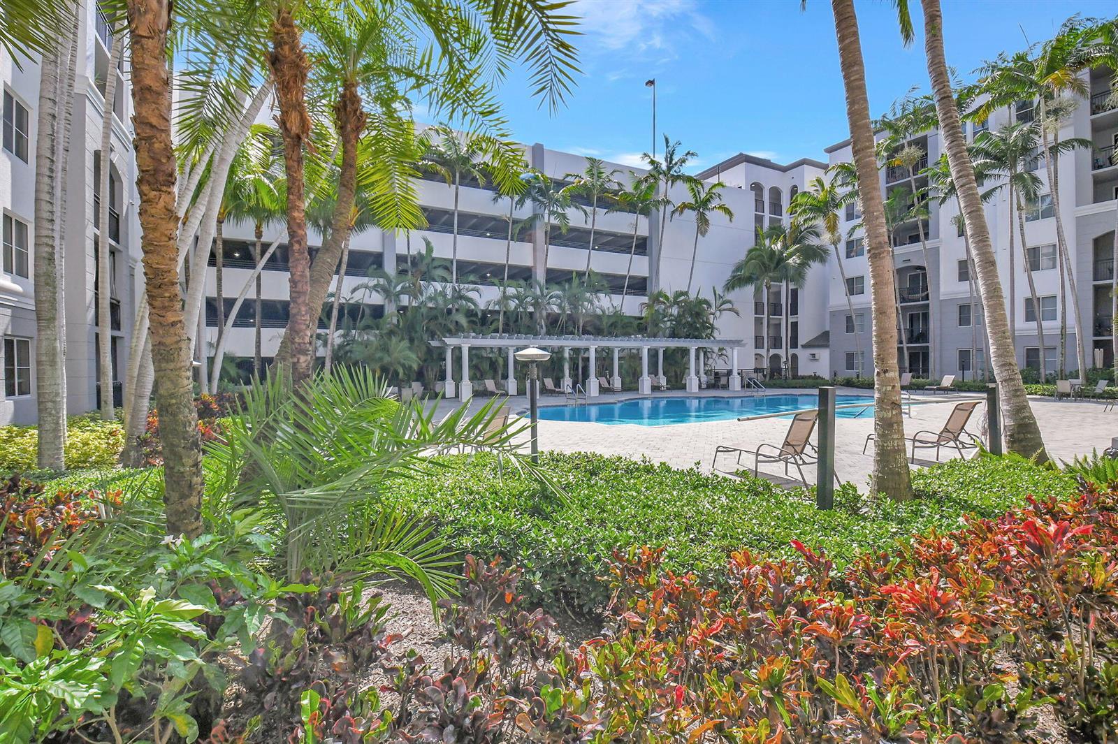 A swimming pool with lounge chairs sits in a courtyard surrounded by palm trees and buildings — 1660 Renaissance Commons Boulevard # 2424, Boynton Beach FL 33426, BOYNTON BEACH, FL
