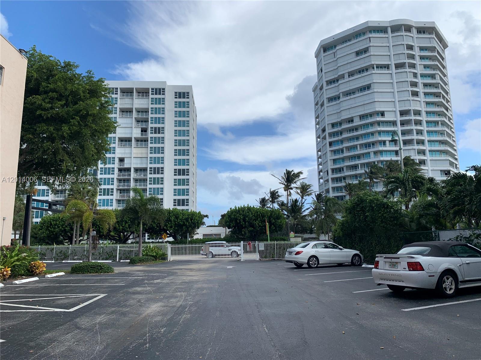 Two tall residential buildings stand behind a parking lot with a white fence — LAUDERDALE BY THE SEA, FL