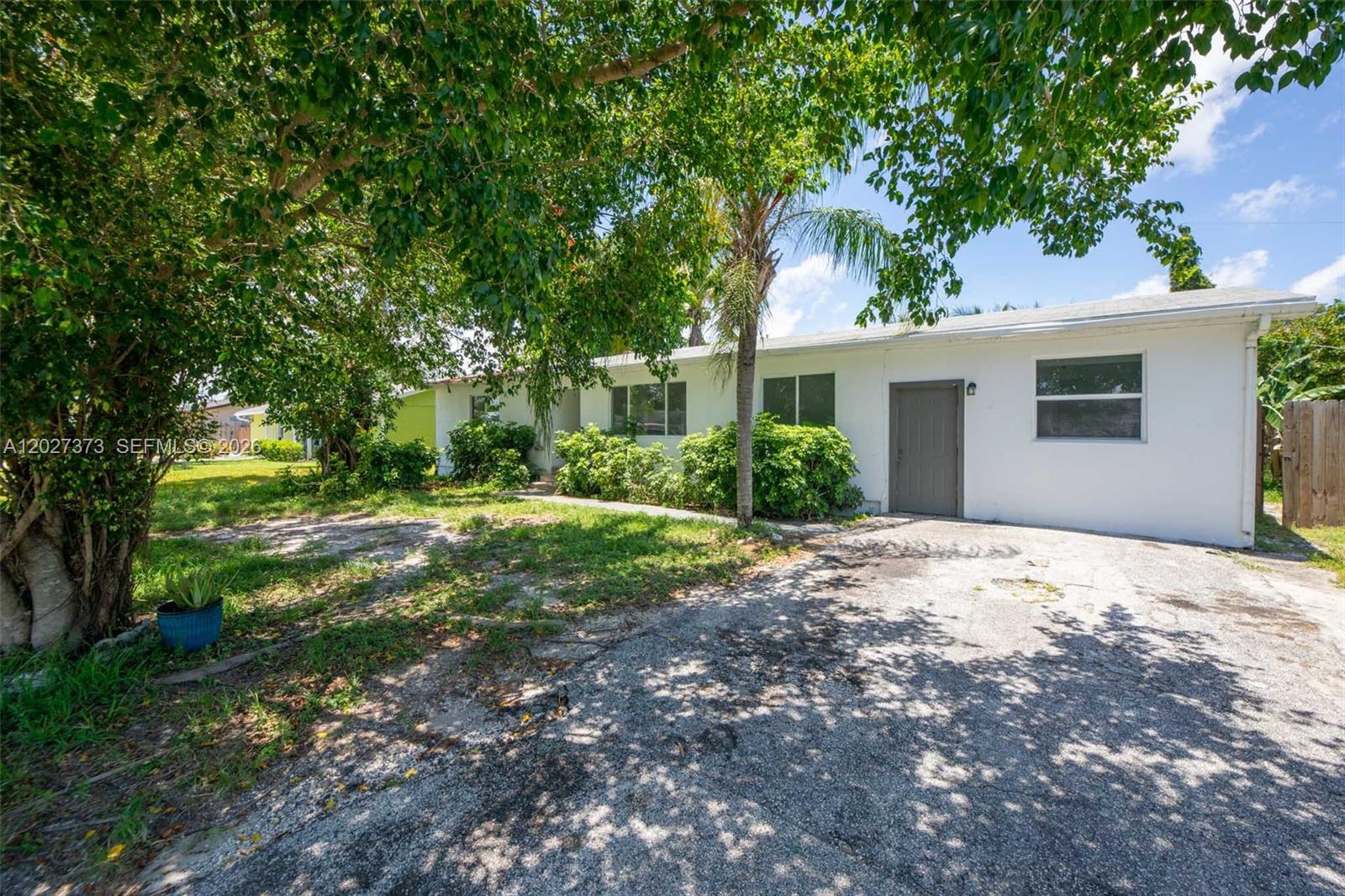A single-story white house features a gravel driveway, mature trees, and a gray front door — 3327 Sapphire Rd, Lake Worth FL 33462, LAKE WORTH, FL