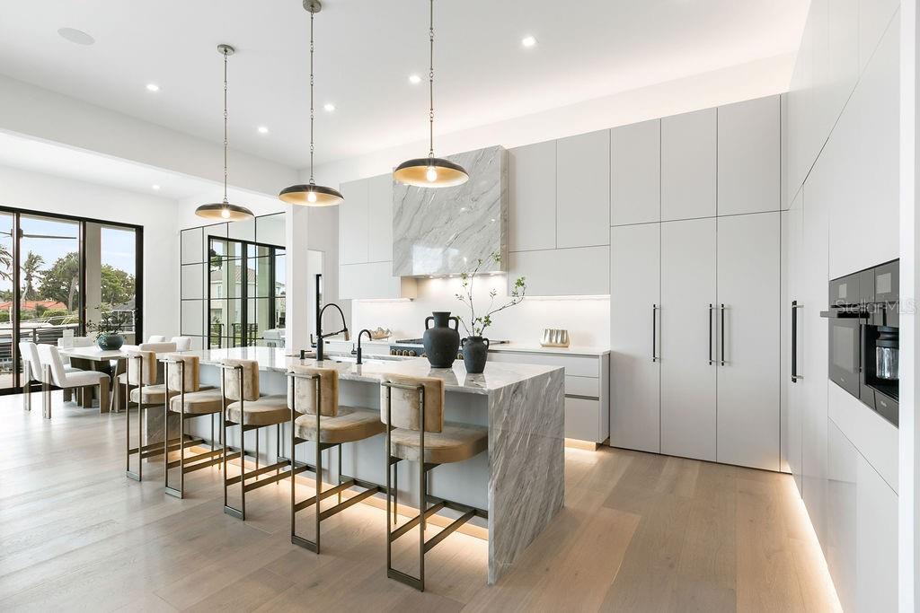 A modern kitchen features a marble waterfall island, light wood floors, and minimalist white cabinetry — 530 CHIPPING LANE, LONGBOAT KEY, FL