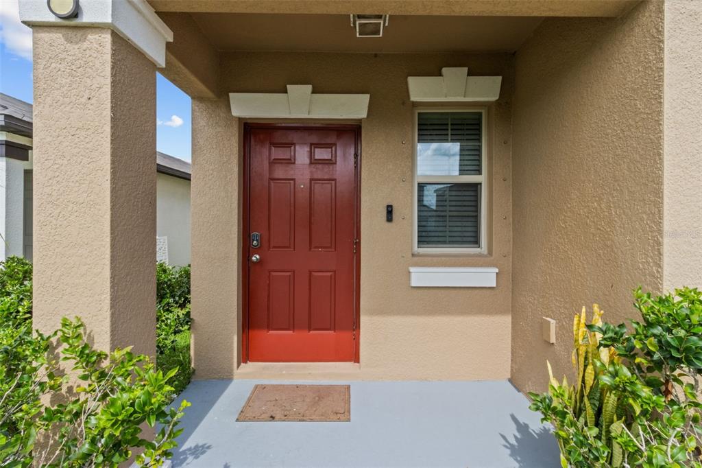 A red front door is centered on a covered porch with a window and plants — 16671 KINGMAN REEF STREET, WIMAUMA, FL