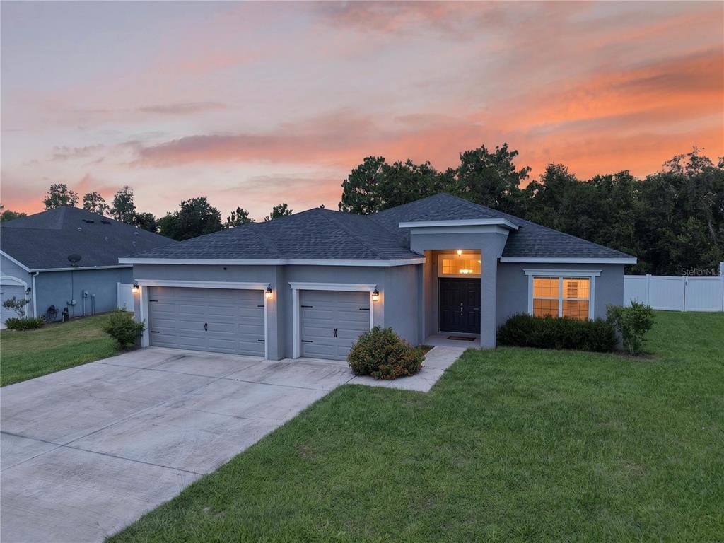 A single-story gray house with a three-car garage and a front walkway at sunset — 11370 SW 57TH AVENUE, OCALA, FL