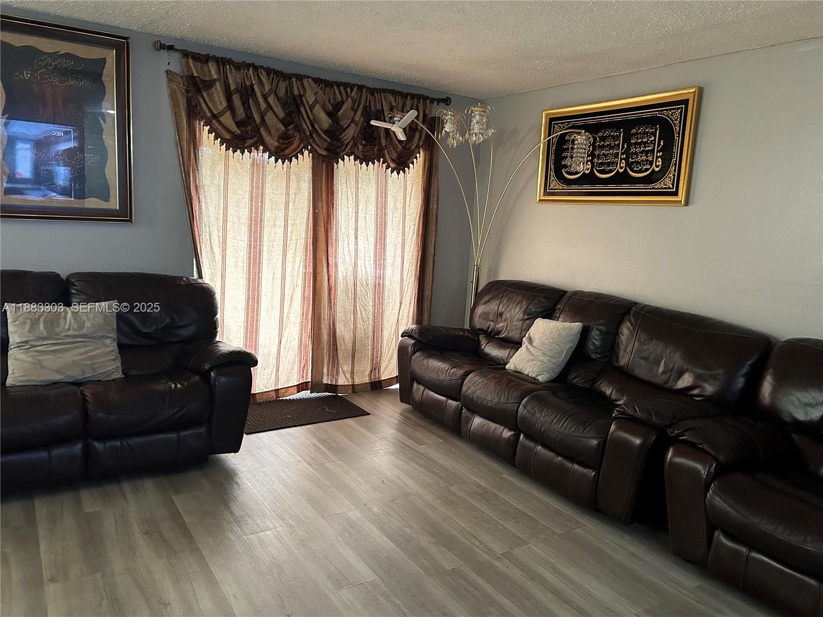 A living room features dark leather sofas, light wood-look flooring, and framed Islamic calligraphy art — 18111 NW 68th Ave # G206, Hialeah FL 33015, HIALEAH, FL
