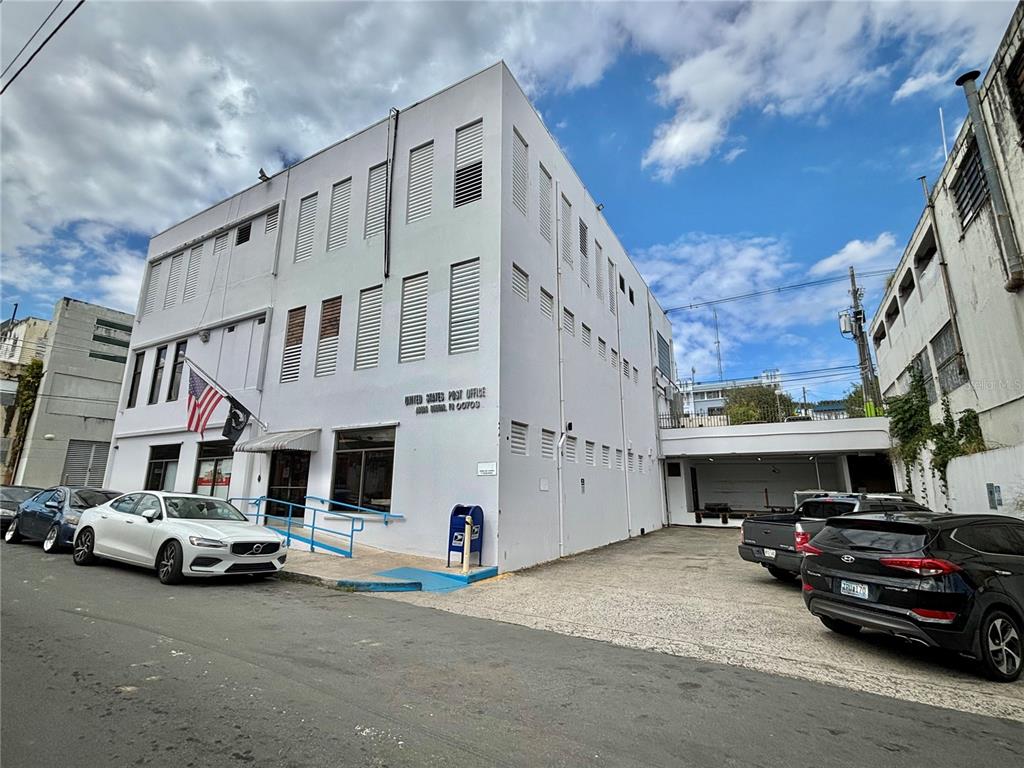 A three-story white building with a post office sign and a blue collection box outside — 20 LUIS MUNOZ RIVERA, AGUAS BUENAS, PR