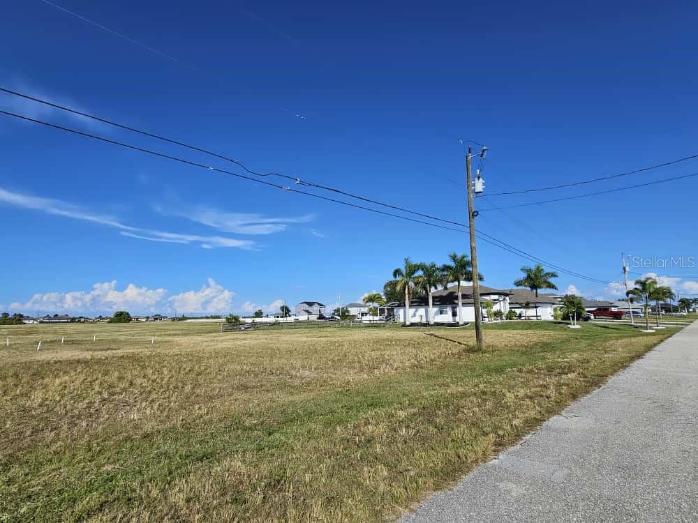 A vacant, grassy lot sits beside a paved road with houses and palm trees nearby — 3405 NW 6TH STREET, CAPE CORAL, FL