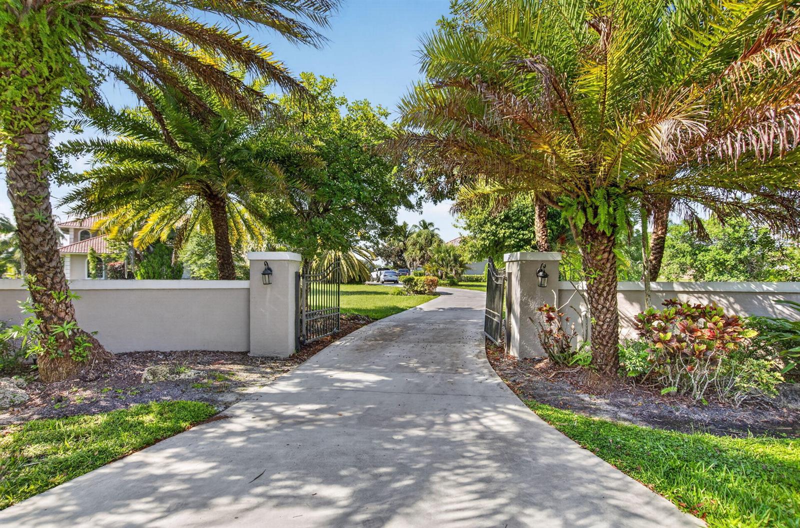A concrete driveway leads through an open metal gate between two stucco pillars and palm trees — 10792 El Caballo Court, Delray Beach FL 33446, DELRAY BEACH, FL