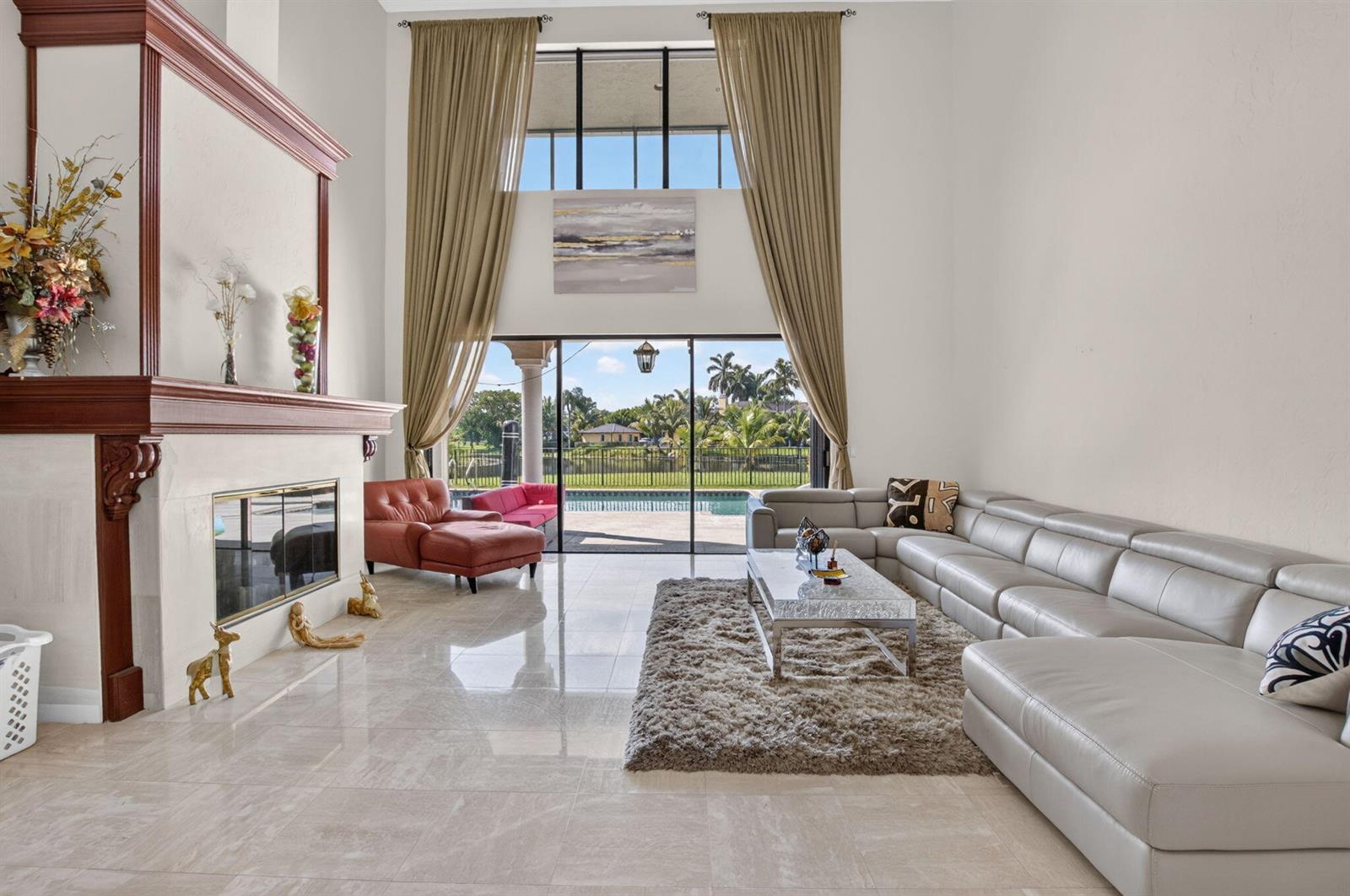 A spacious living room features a fireplace, tall windows, and a view of a pool — 10792 El Caballo Court, Delray Beach FL 33446, DELRAY BEACH, FL