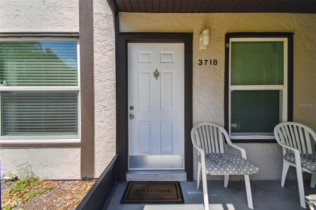 A white front door with a metal kickplate sits between two windows and two chairs — 3718 59TH AVE W UNIT 3718, BRADENTON, FL