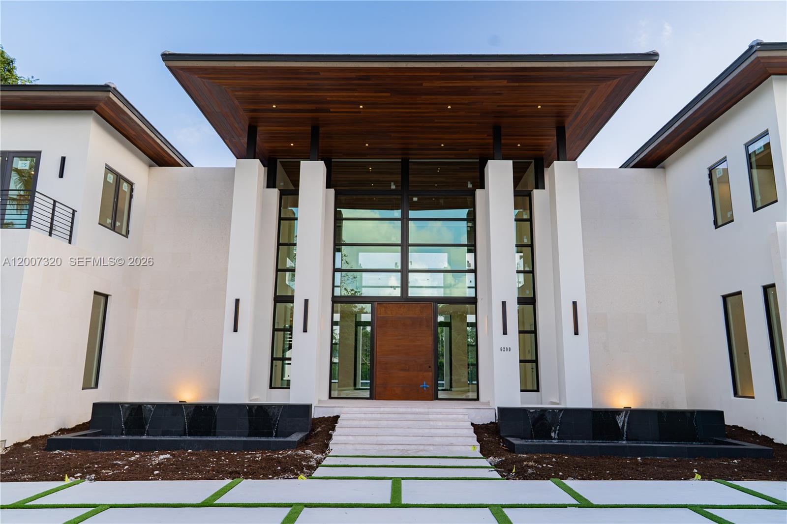 The exterior of a modern house features a large wooden overhang, glass entryway, and fountains — 6290 Chapman Field Dr, Pinecrest FL 33156, PINECREST, FL