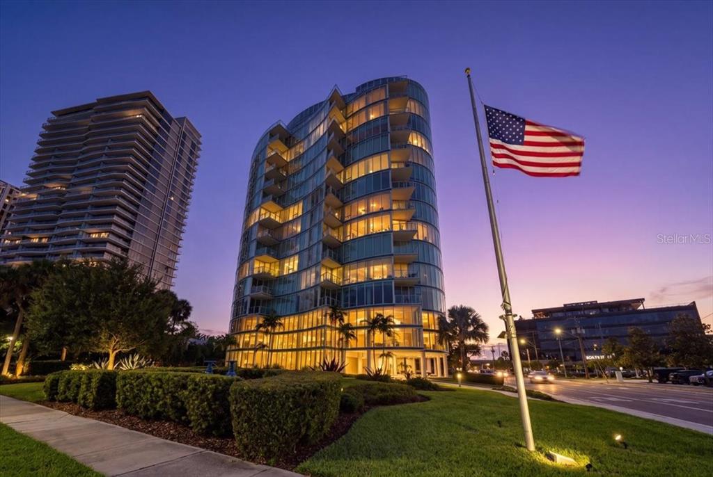 A modern glass-walled residential building stands at dusk next to a tall American flag — 2900 W BAY TO BAY BOULEVARD UNIT 1401, TAMPA, FL