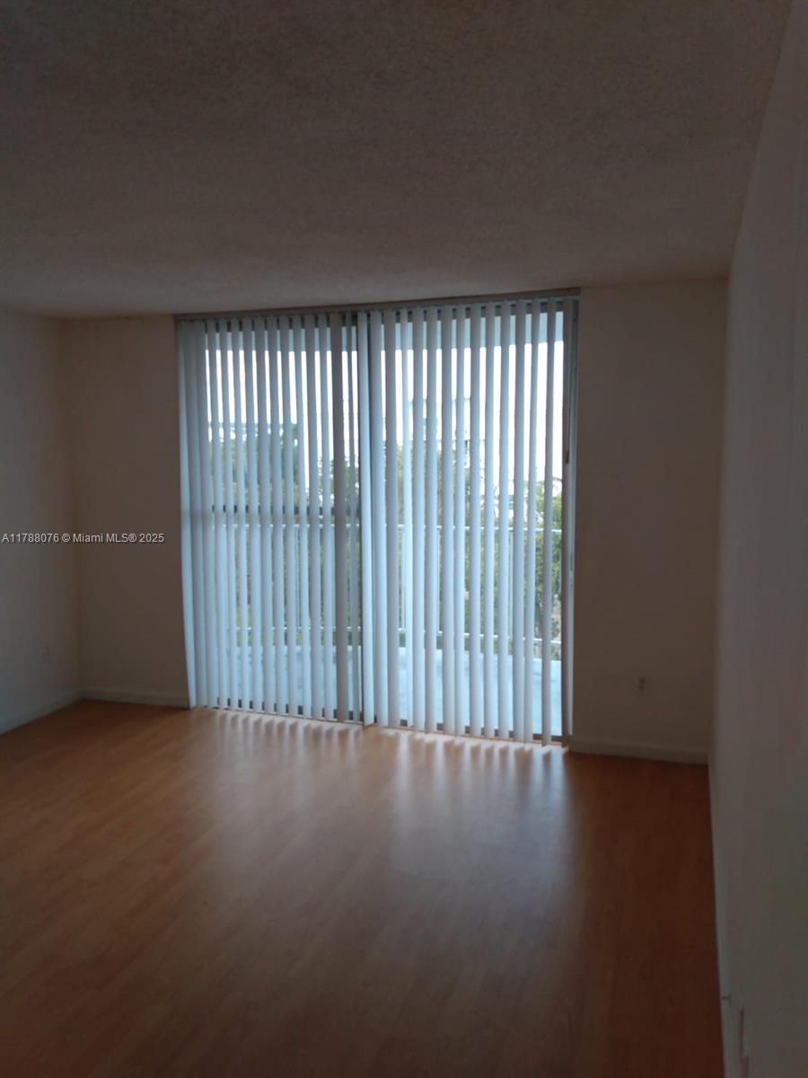 An empty room features light wood-look flooring and a large sliding glass door with vertical blinds — 494 NW 165th St Rd # 102, Miami FL 33169, MIAMI, FL