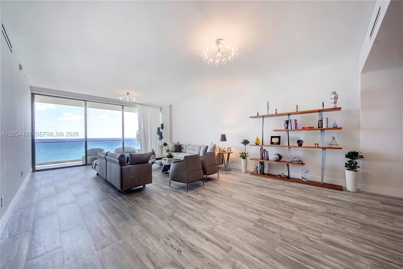 A modern living room features wood-look tile flooring, a large bookshelf, and floor-to-ceiling ocean views — 10203 Collins Ave # 1502, Bal Harbour FL 33154, BAL HARBOUR, FL