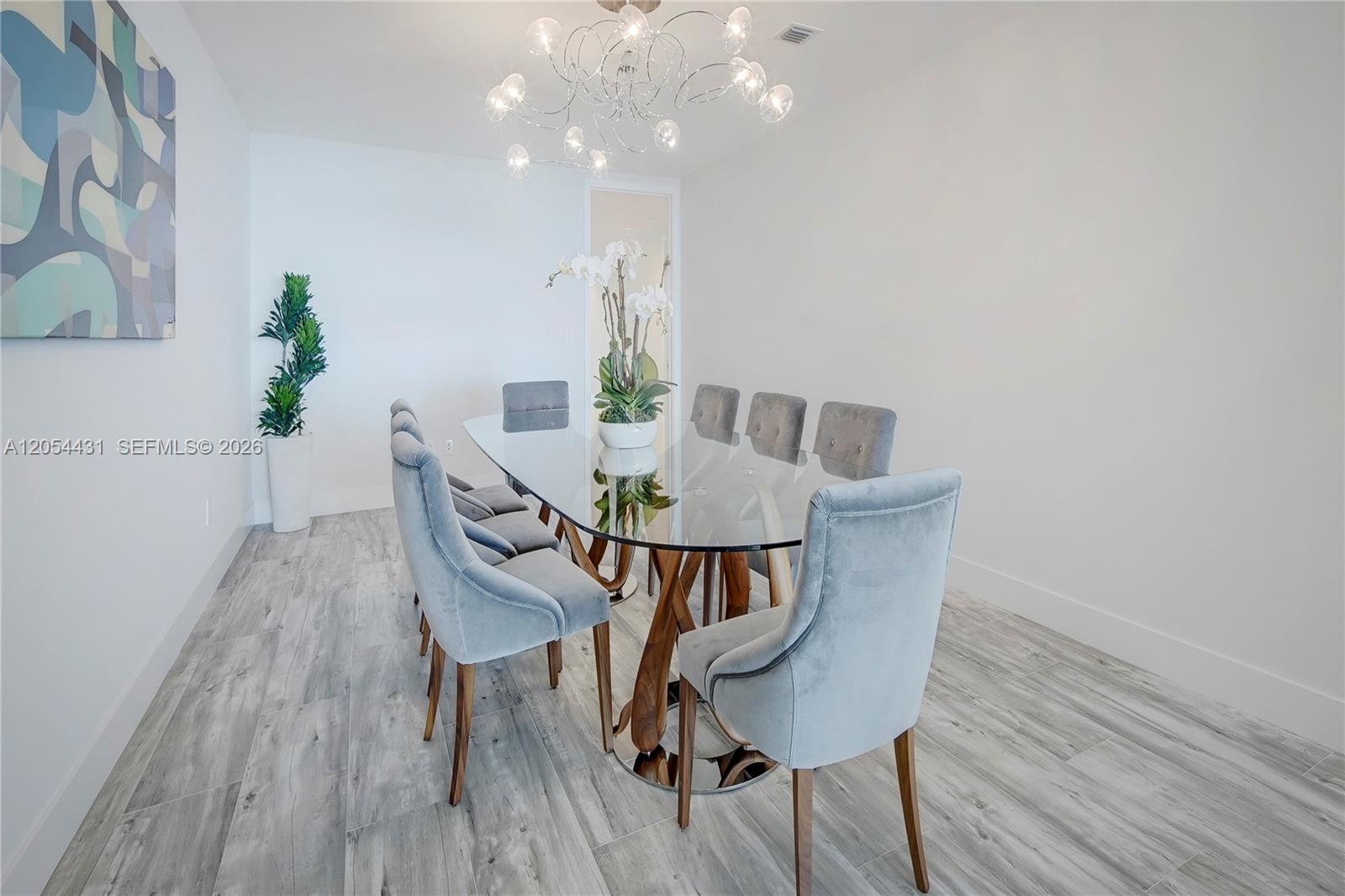 A dining room features a glass-topped table, gray velvet chairs, and a modern chandelier — 10203 Collins Ave # 1502, Bal Harbour FL 33154, BAL HARBOUR, FL