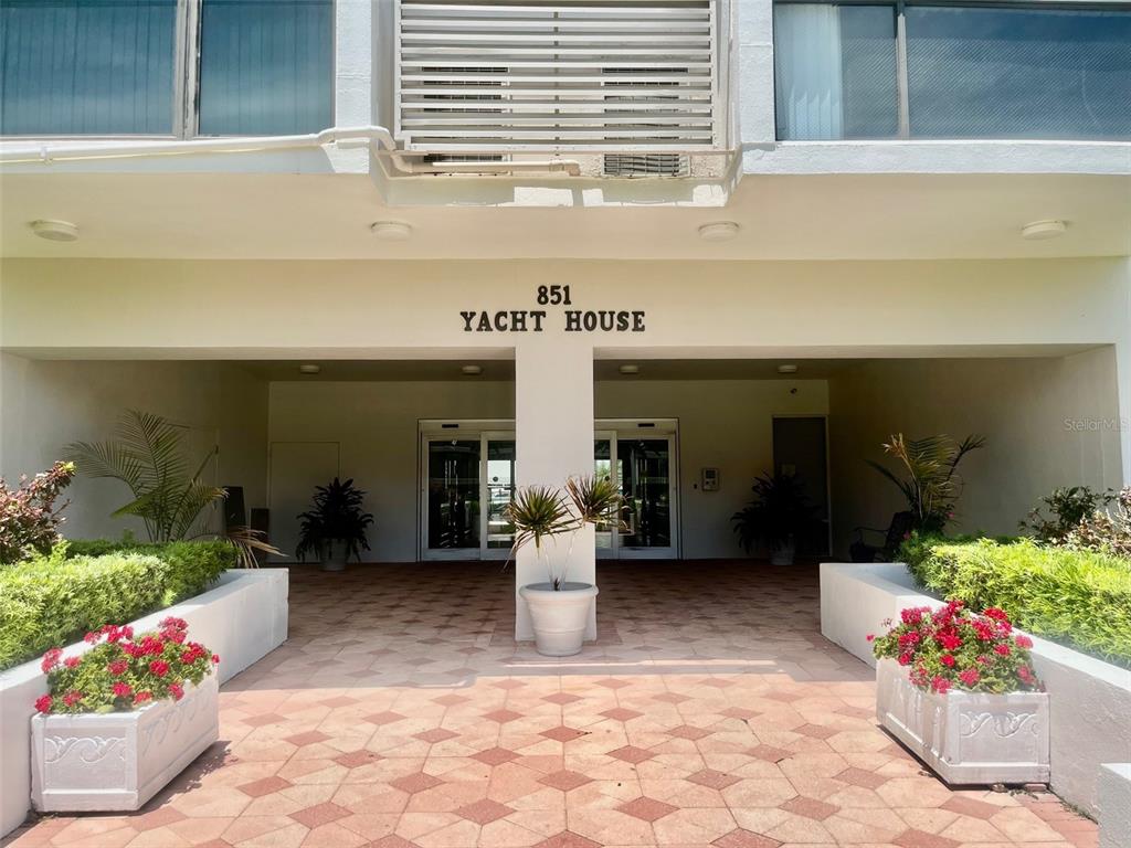 The exterior entrance of a building features a paved walkway, potted plants, and red flowers — 851 BAYWAY BOULEVARD UNIT 405, CLEARWATER BEACH, FL