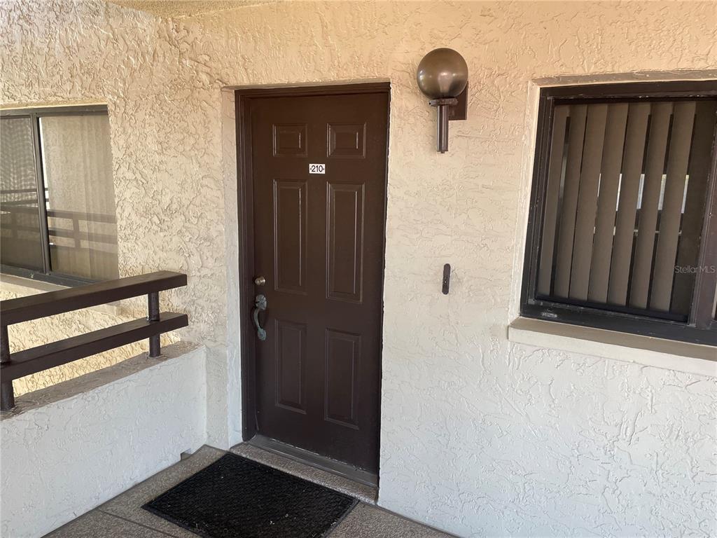 A brown front door with the number 210 is set in a textured stucco wall — 106 1ST STREET E UNIT 210, TIERRA VERDE, FL