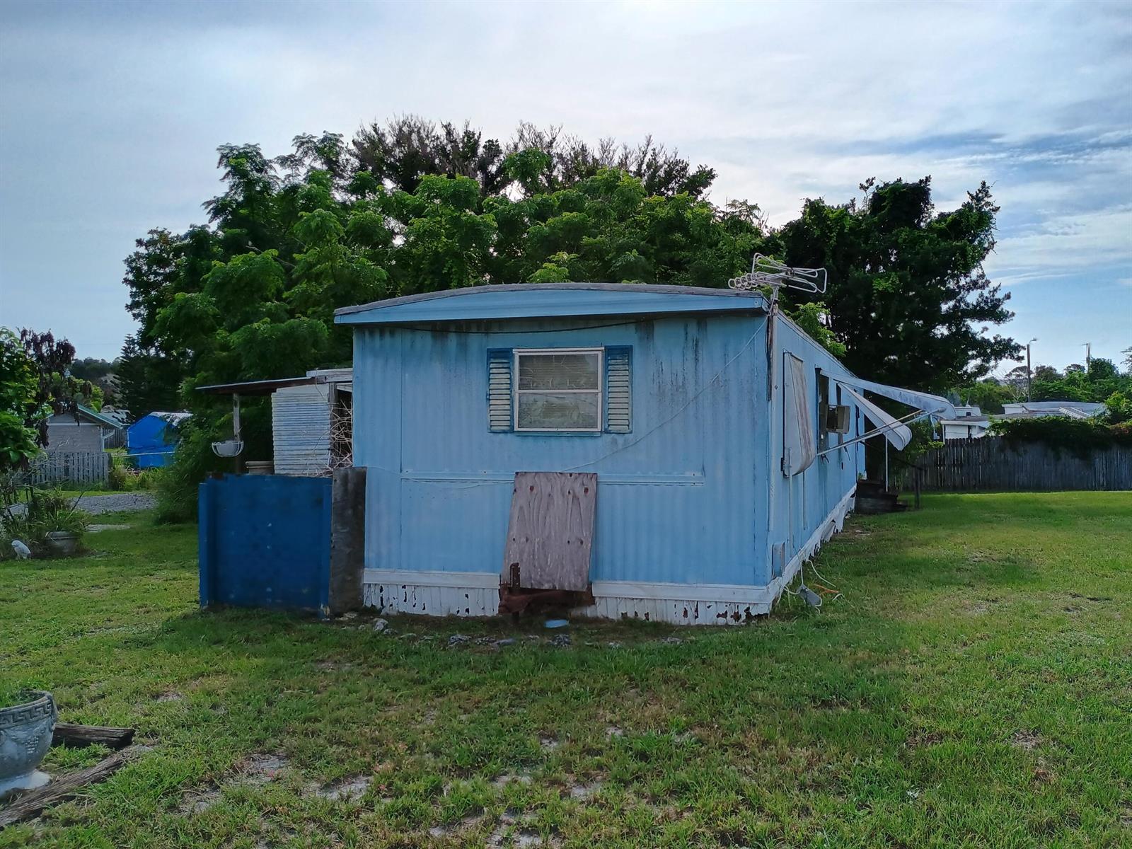 A blue mobile home with a plywood-covered section sits on a grassy lot — 1512 NE Dove Avenue, Stuart FL 34994, STUART, FL