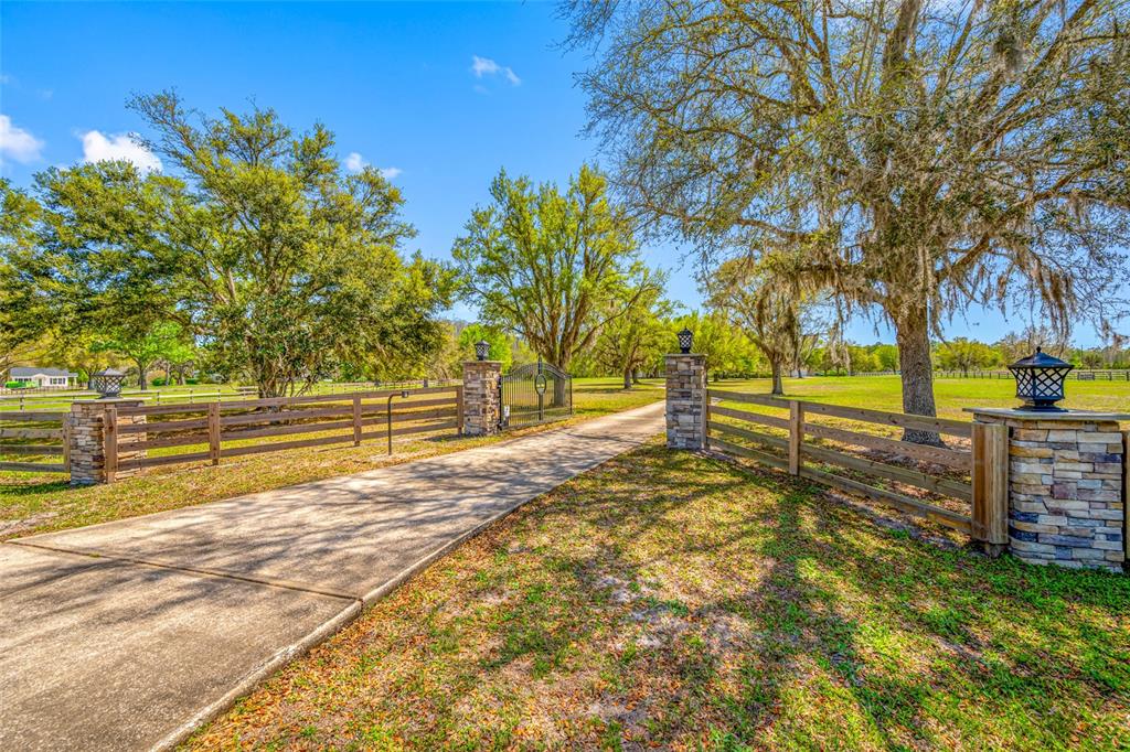A concrete driveway leads through a gated entrance flanked by stone pillars and wooden fencing — 11939 PASCO TRAILS BOULEVARD, SPRING HILL, FL
