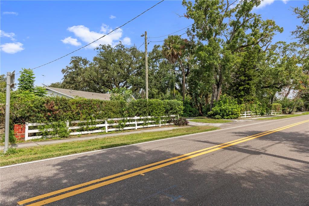 A white fence borders a grassy yard with mature trees along a paved road — 6401 WINGFOOT CIRCLE, TAMPA, FL