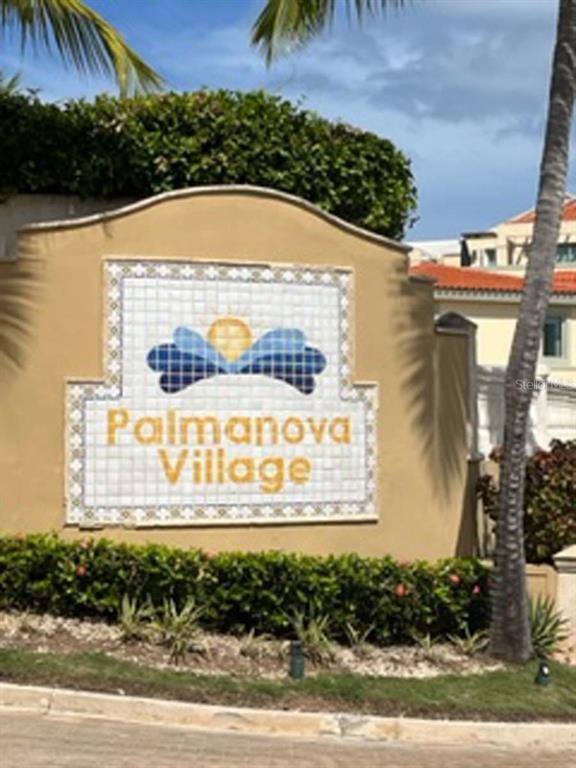 A tan stucco sign with a tiled logo reads Palmanova Village in front of a building — Palms Inn Drive PALMANOVA VILLAGE UNIT 2-304, HUMACAO, PR