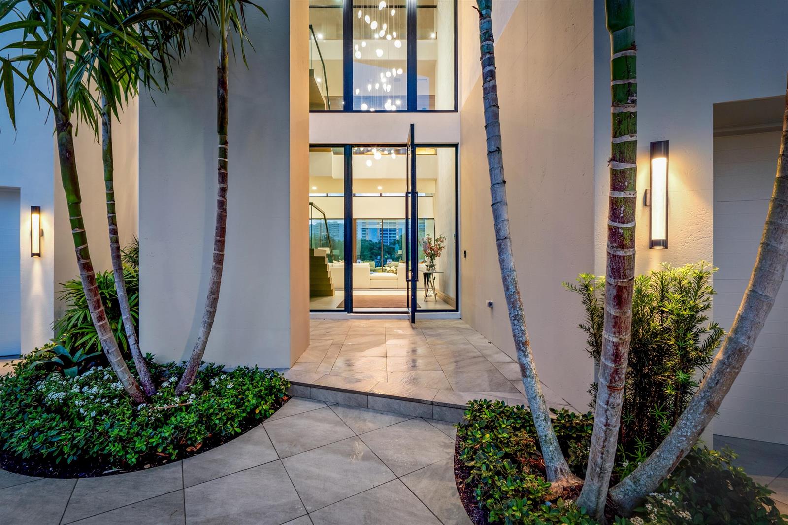 A modern home entrance features a tall glass pivot door, palm trees, and outdoor lighting — 4080 Ibis Point Circle, Boca Raton FL 33431, BOCA RATON, FL