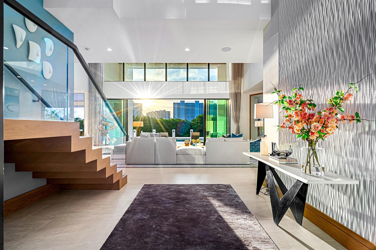 A modern living room with a floating staircase, textured wall, and floor-to-ceiling windows overlooking buildings — 4080 Ibis Point Circle, Boca Raton FL 33431, BOCA RATON, FL