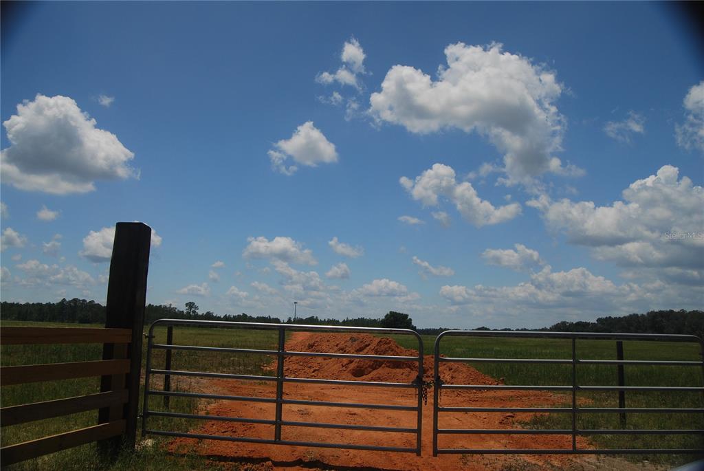 A metal gate opens to a dirt path leading through a grassy field under clouds — LAKE ERIE, GROVELAND, FL