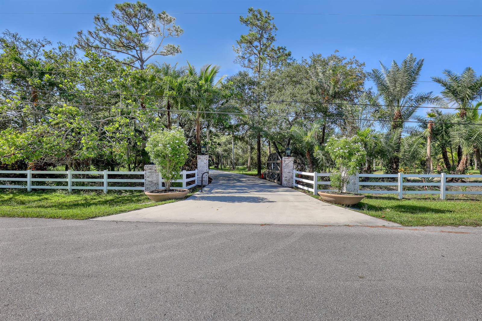 A concrete driveway leads through a gated entrance flanked by white fences and potted trees — 10780 Anderson Lane, Wellington FL 33449, WELLINGTON, FL
