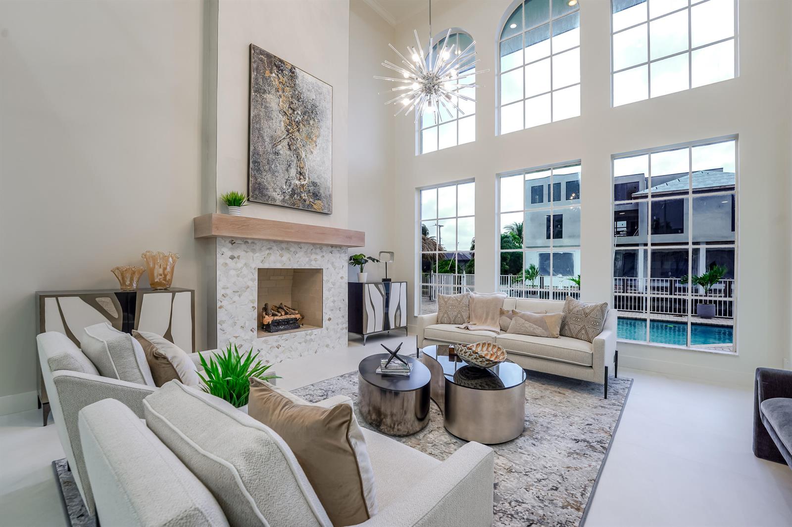 A bright living room features a tall fireplace, modern chandelier, and large windows overlooking a pool — 749 NE 71st Street, Boca Raton FL 33487, BOCA RATON, FL