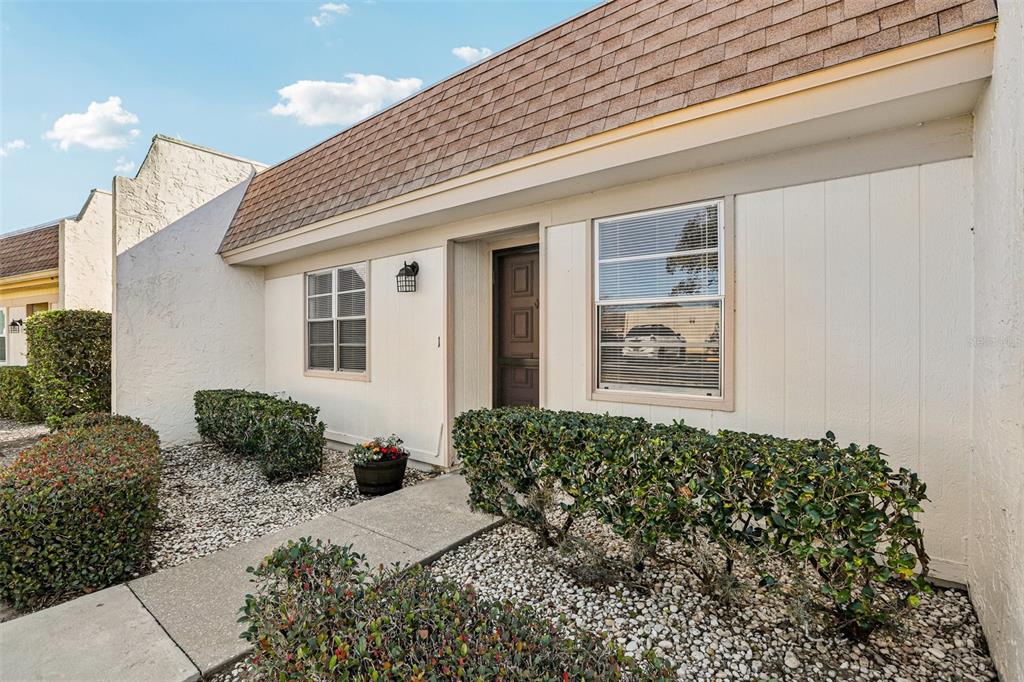 The exterior of a one-story home features white siding, a brown shingled roof, and landscaping — 11120 CARRIAGE HILL DRIVE UNIT 1, PORT RICHEY, FL