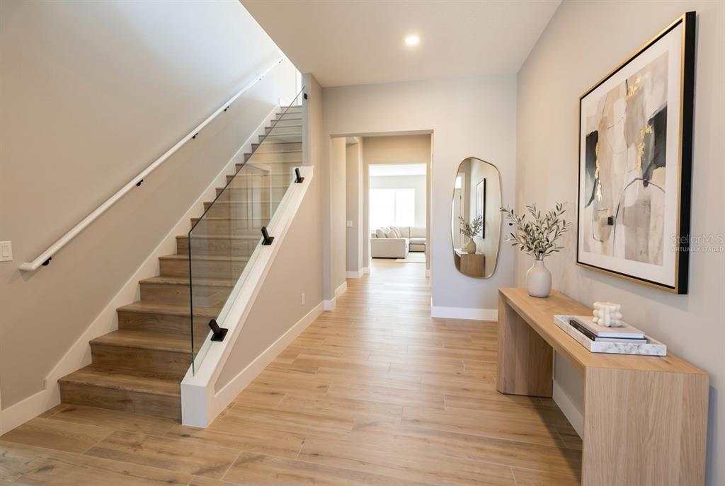 A modern entryway features wood-look tile flooring, a glass-railed staircase, and a console table — 16046 VETTA DRIVE, BELLA COLLINA, FL