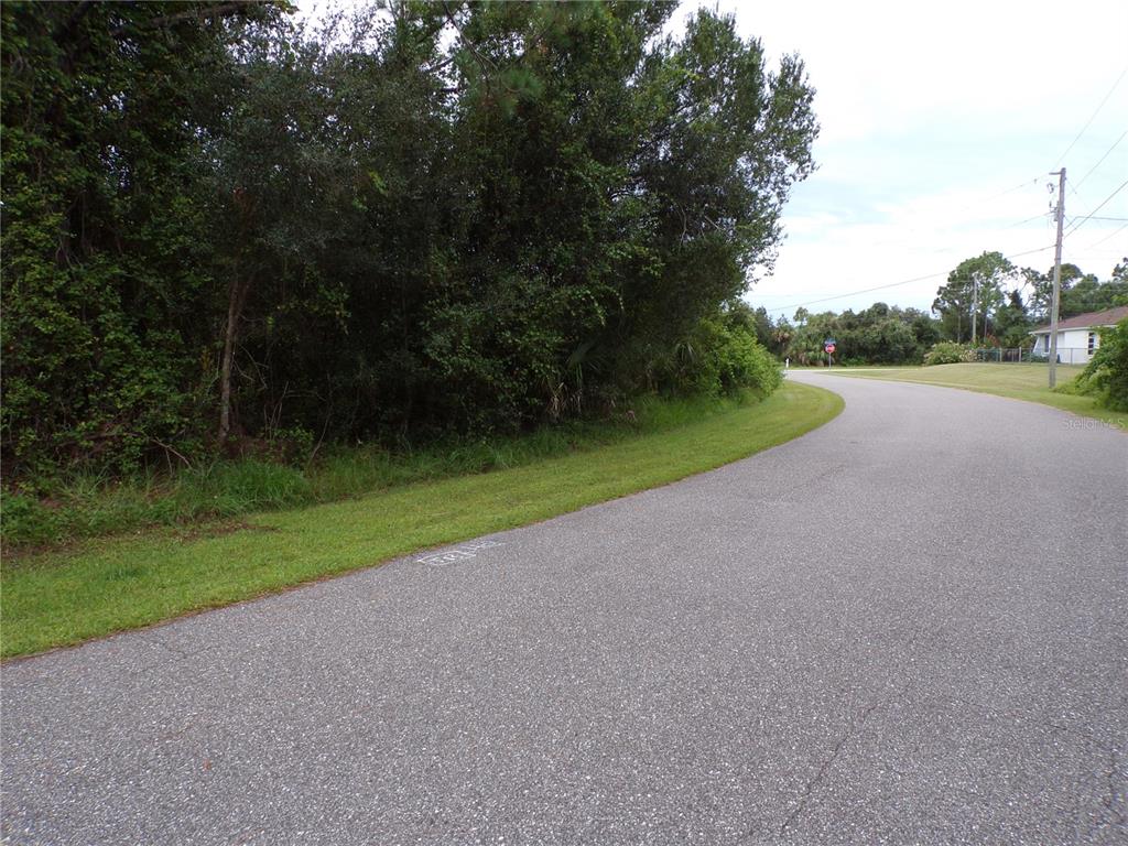 A paved road curves past a grassy shoulder and a dense area of trees — EMBASSY ROAD, NORTH PORT, FL